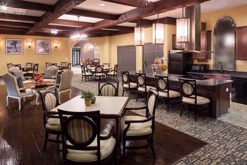 A spacious, warmly lit common area with a combination of dining and lounge seating. The room features multiple tables with chairs, a kitchen island with bar stools, pendant lighting, and exposed wooden ceiling beams. The walls are painted yellow and decorated with framed black and white photographs.