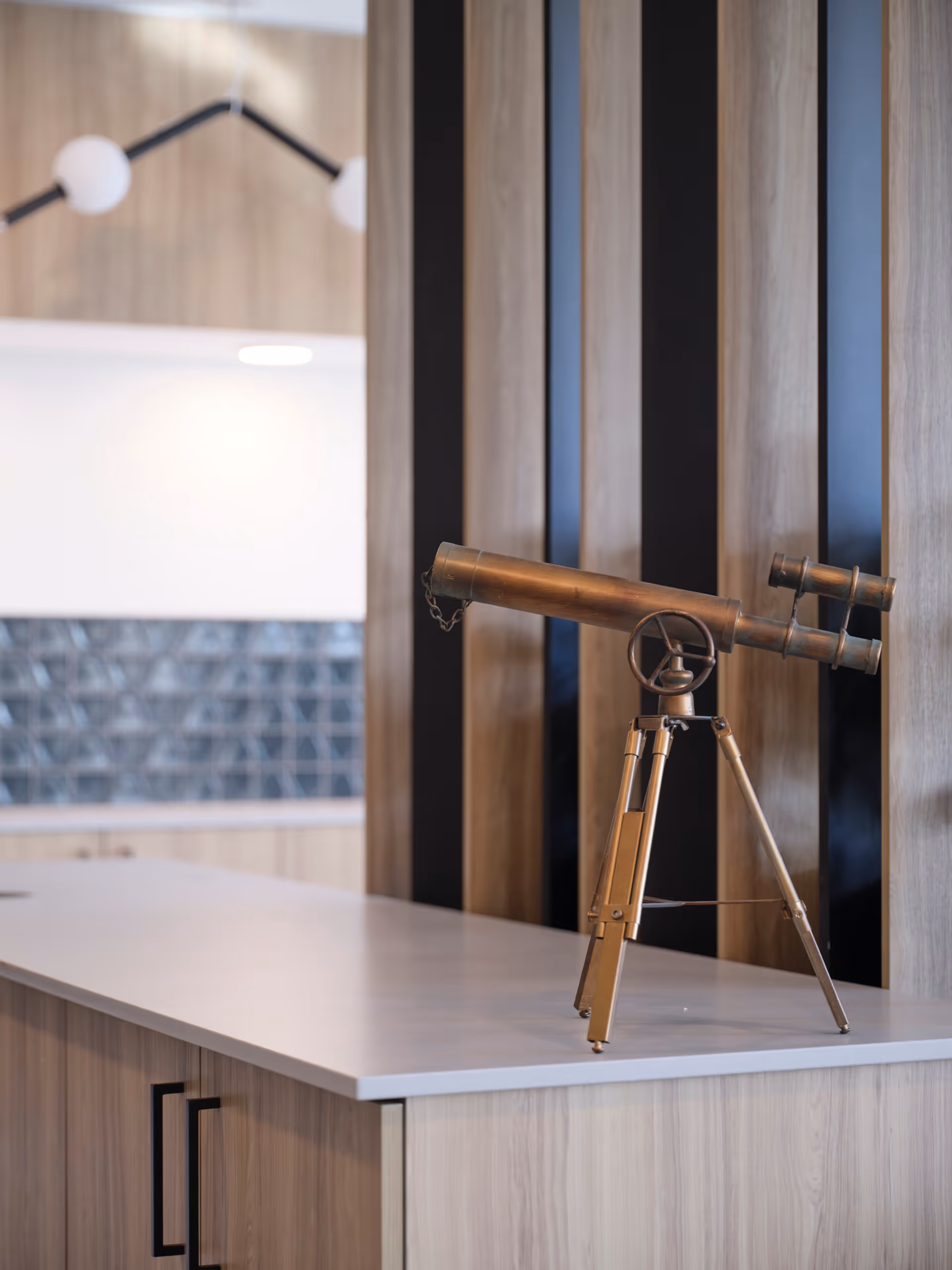 A vintage brass telescope mounted on a tripod stands on a light wood countertop in a modern interior space with vertical wood and black paneling in the background.