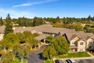 Aerial view of Dale Commons senior living facility surrounded by trees and greenery under a clear blue sky, showing the building's roof, entrance, and adjacent parking area.
