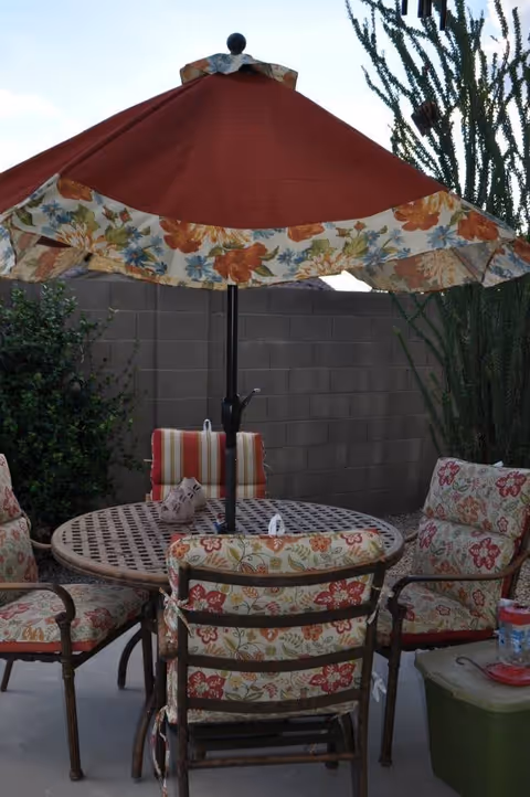 Outdoor patio area with a round metal table and four cushioned chairs featuring floral and striped patterns. A large red umbrella with a floral trim is centered over the table. The setting is enclosed by a brick wall with some greenery and tall plants in the background.