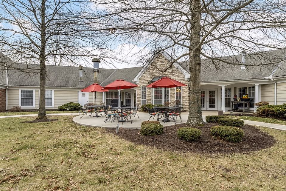 Outdoor courtyard with round tables, red umbrellas, and landscaping in front of a single-story senior living building.