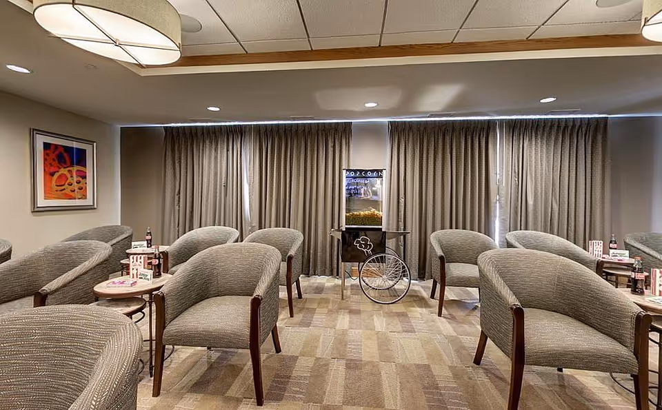 A cozy lounge area with multiple gray upholstered armchairs arranged in rows facing a popcorn machine. Small round tables between the chairs hold popcorn boxes and bottles of soda. The room has beige walls, a patterned carpet, a framed abstract painting on the left wall, and large windows covered with beige curtains.