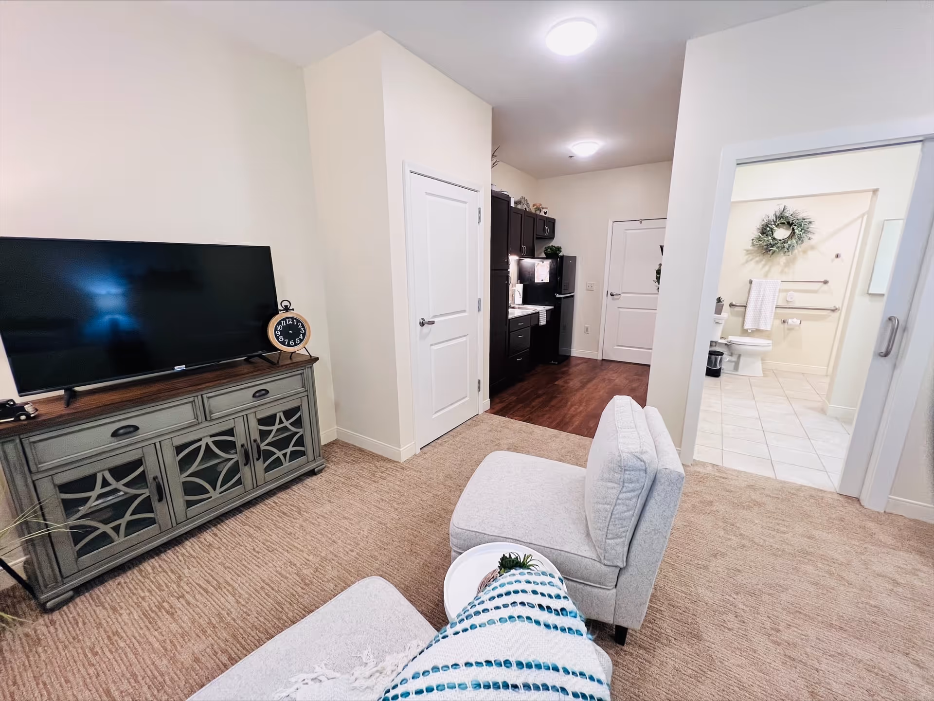 Interior view of a senior living facility room showing a living area with a TV on a cabinet, a light gray chair with a blue and white blanket, a small kitchen area with dark cabinets and appliances, and a bathroom with a toilet and towel rack visible through an open door.
