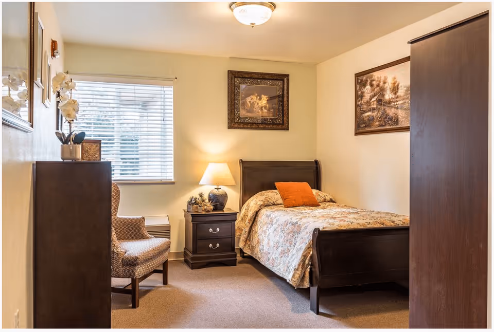 A cozy bedroom in a senior living memory care facility featuring a single bed with floral bedding and an orange pillow, a wooden nightstand with a lamp and decorative items, a patterned armchair, a window with blinds, and framed artwork on the walls.