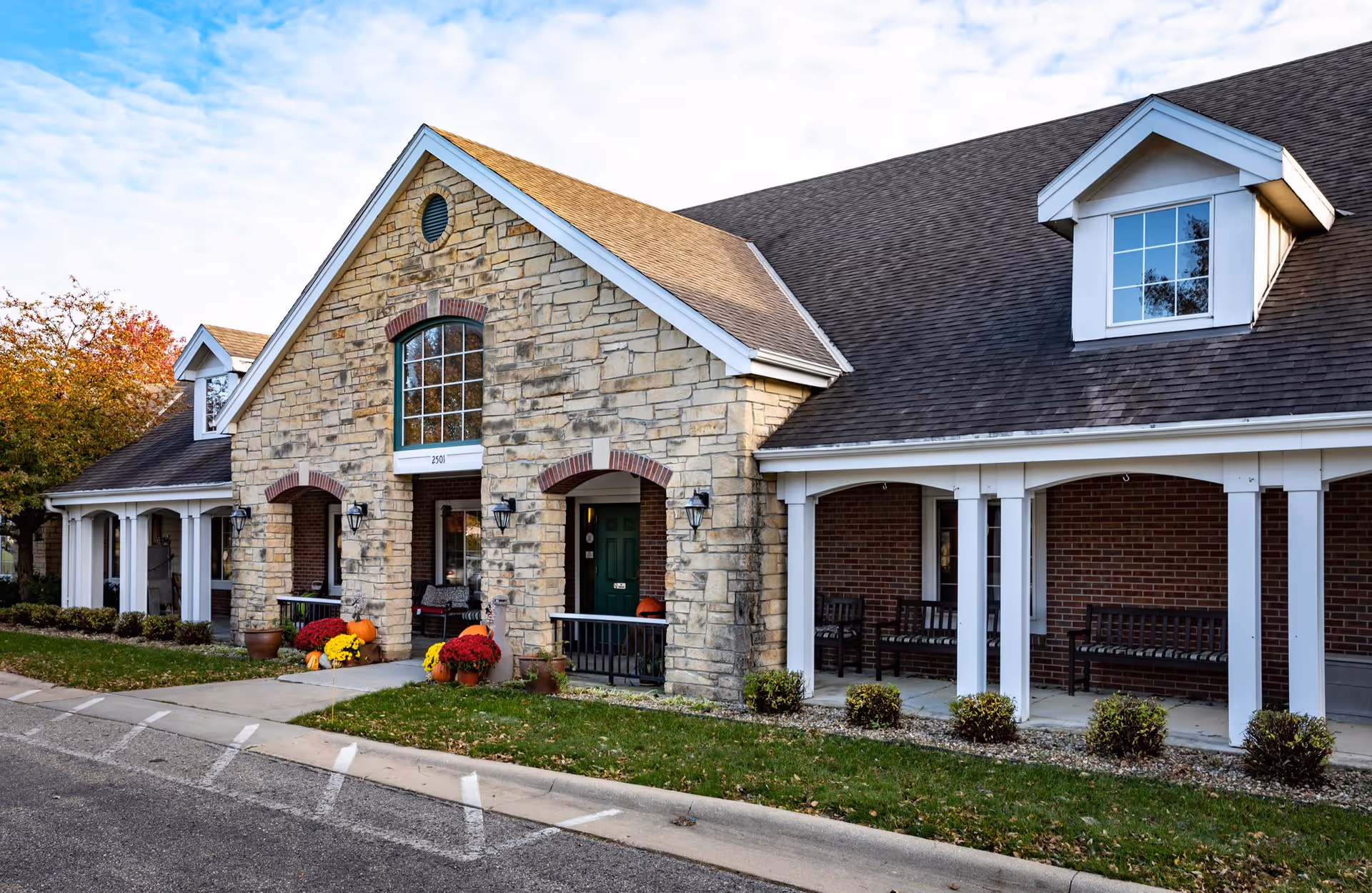 Exterior view of a senior living facility building with stone and brick facade, a green door, large windows, and a covered porch with benches. There are potted plants and colorful flowers near the entrance, and a paved driveway in front.