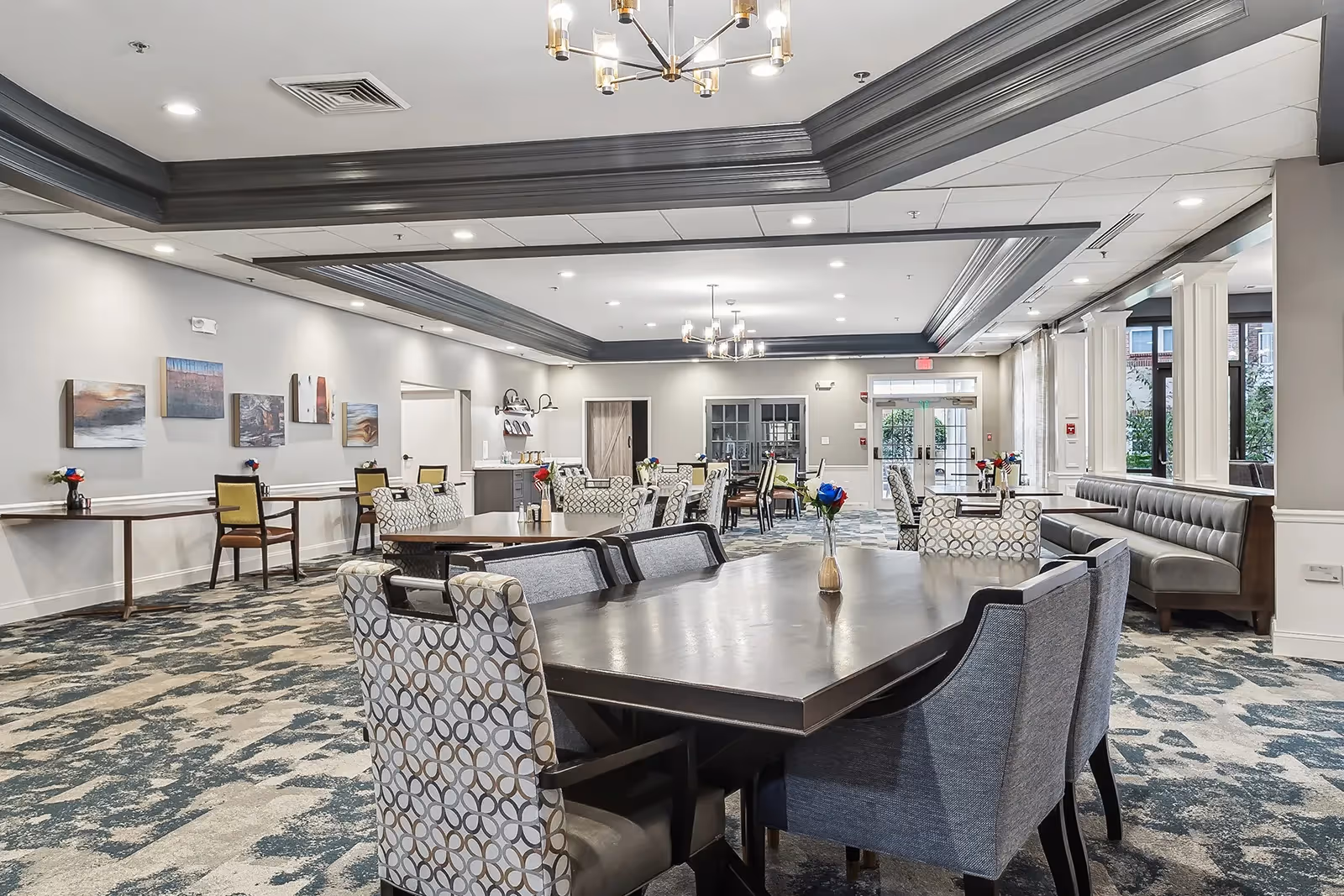Open, well-lit communal dining and lounge area with multiple tables, upholstered chairs, and chandeliers.