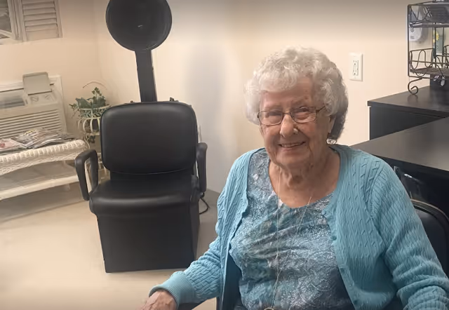 An elderly woman with white curly hair and glasses is smiling while sitting in a chair inside a room. She is wearing a blue patterned top and a light blue cardigan. Behind her, there is a black salon chair with a hair dryer hood, a white wicker bench with magazines, and a small plant on a stand.