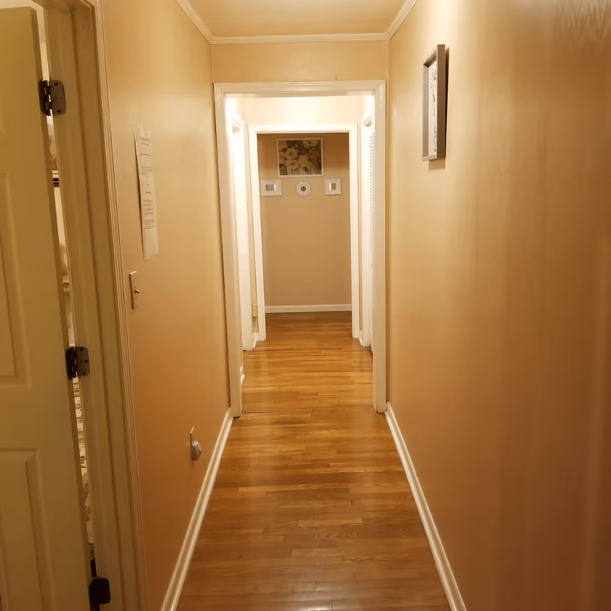 A narrow hallway with wooden flooring and beige walls. There are white door frames on both sides of the hallway and a few framed pictures hanging on the walls. The hallway is well-lit with ceiling lighting.