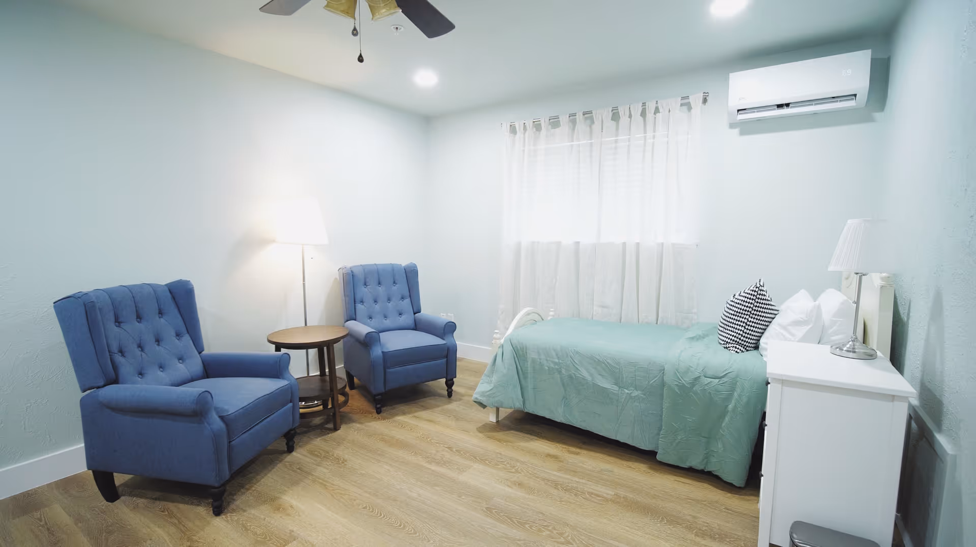 A bright, clean bedroom with light blue walls and wood flooring. The room features a single bed with a green bedspread and multiple pillows, a white nightstand with a lamp, two blue upholstered armchairs, a small round wooden table between the chairs, a floor lamp, a ceiling fan, and a window with sheer white curtains. An air conditioning unit is mounted on the wall above the bed.