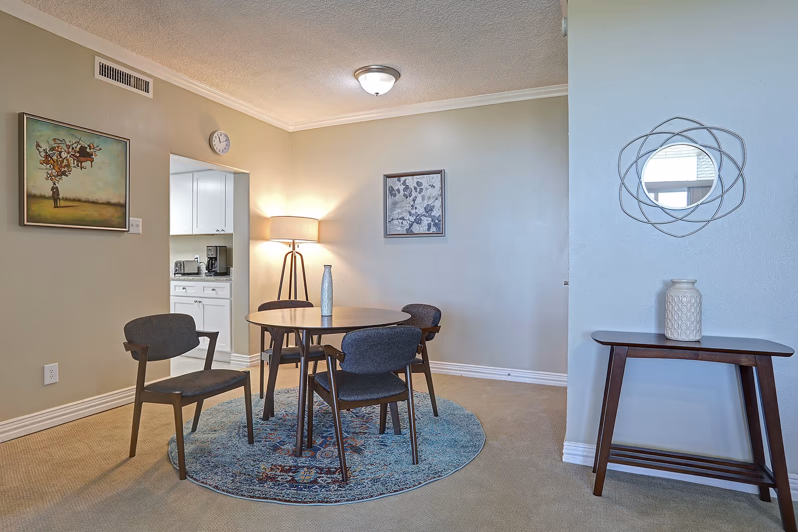 A small dining area with a round wooden table and four dark cushioned chairs on a circular patterned rug. A floor lamp is lit in the corner, and two framed artworks hang on the beige walls. To the right, there is a wooden console table with a decorative vase and a round mirror with a geometric frame above it. A kitchen with white cabinets and appliances is visible through an open doorway.