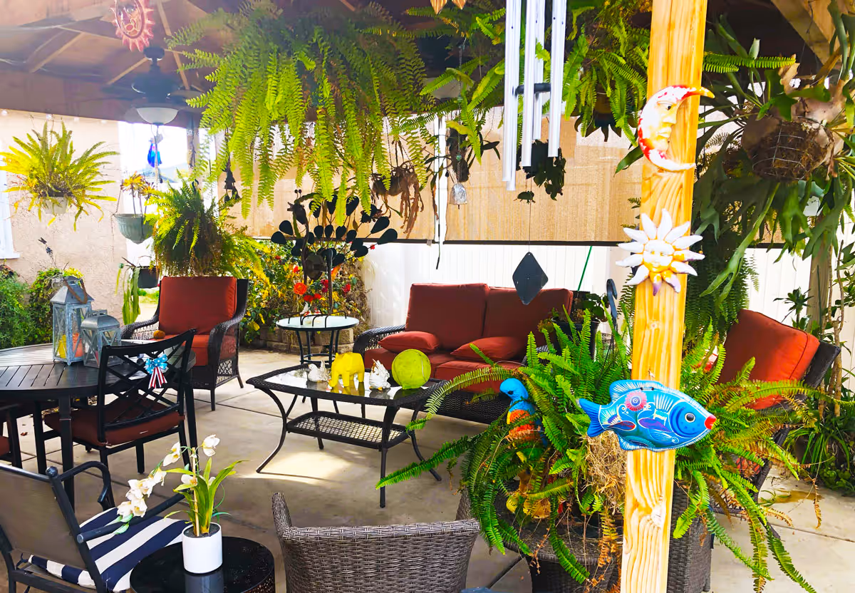 Covered outdoor patio area with various seating including cushioned chairs and a sofa, surrounded by numerous hanging and potted green plants. Decorative items such as a sun, moon, and fish are attached to a wooden post, and wind chimes hang from the ceiling. The space is bright and inviting with a concrete floor and a table with lanterns and small decorative figurines.