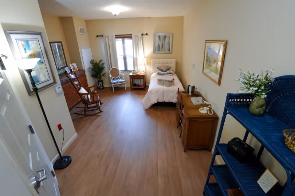 A cozy bedroom in a senior living facility featuring a single bed with pillows and a blanket, a wooden rocking chair, a small side table with a lamp, a window with white curtains, framed artwork on the walls, a wooden dresser, and a blue wicker shelving unit with decorative items and flowers.