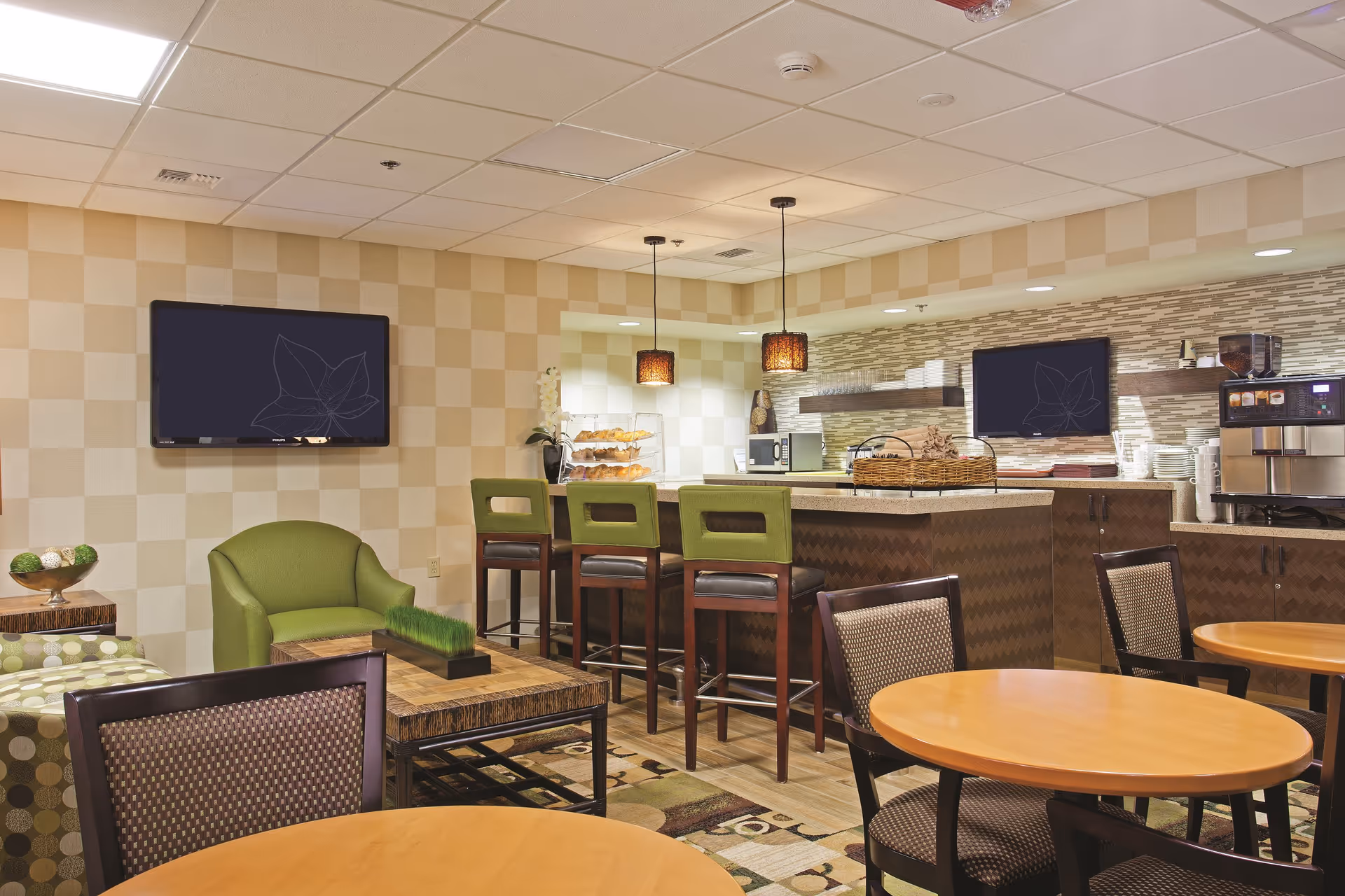 Communal dining and lounge area with round wooden tables, chairs, green barstools at a serving counter, TVs, and a coffee station.