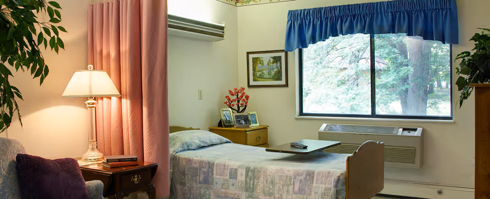 A single bed with a patterned bedspread in a nursing facility room. Next to the bed is a wooden nightstand with framed photos and a decorative plant. A window with blue valance shows trees outside. A lamp on a small table beside a chair with a purple pillow is also visible. The room has light-colored walls and an air conditioning unit below the window.