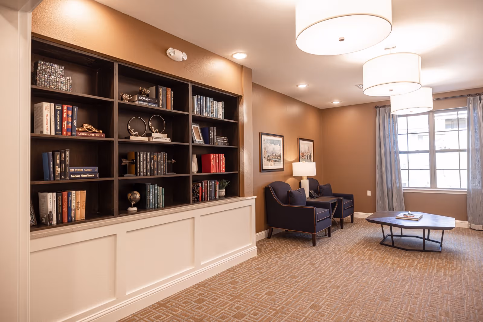 A cozy senior living common area with a built-in dark wood bookshelf filled with books and decorative items on the left. Two navy blue armchairs with a small side table and lamp are positioned against a brown wall with framed artwork. A large window with light curtains lets in natural light, and a modern hexagonal coffee table sits on a patterned carpet under three large ceiling lights.