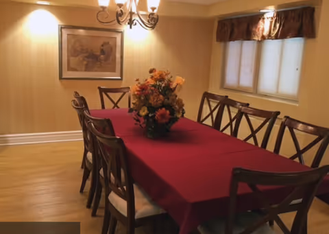 Dining room with a long table covered in a red tablecloth, a floral centerpiece, multiple wooden chairs, a chandelier, and framed art on the wall.