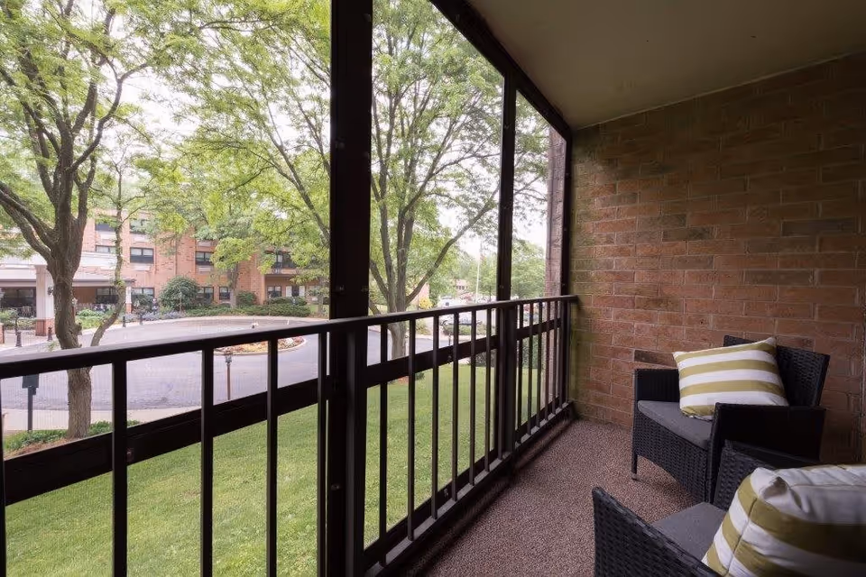 A screened balcony with two black wicker chairs featuring gray cushions and striped yellow and white pillows. The balcony overlooks a green lawn and trees, with a view of a brick building and a circular driveway in the background.