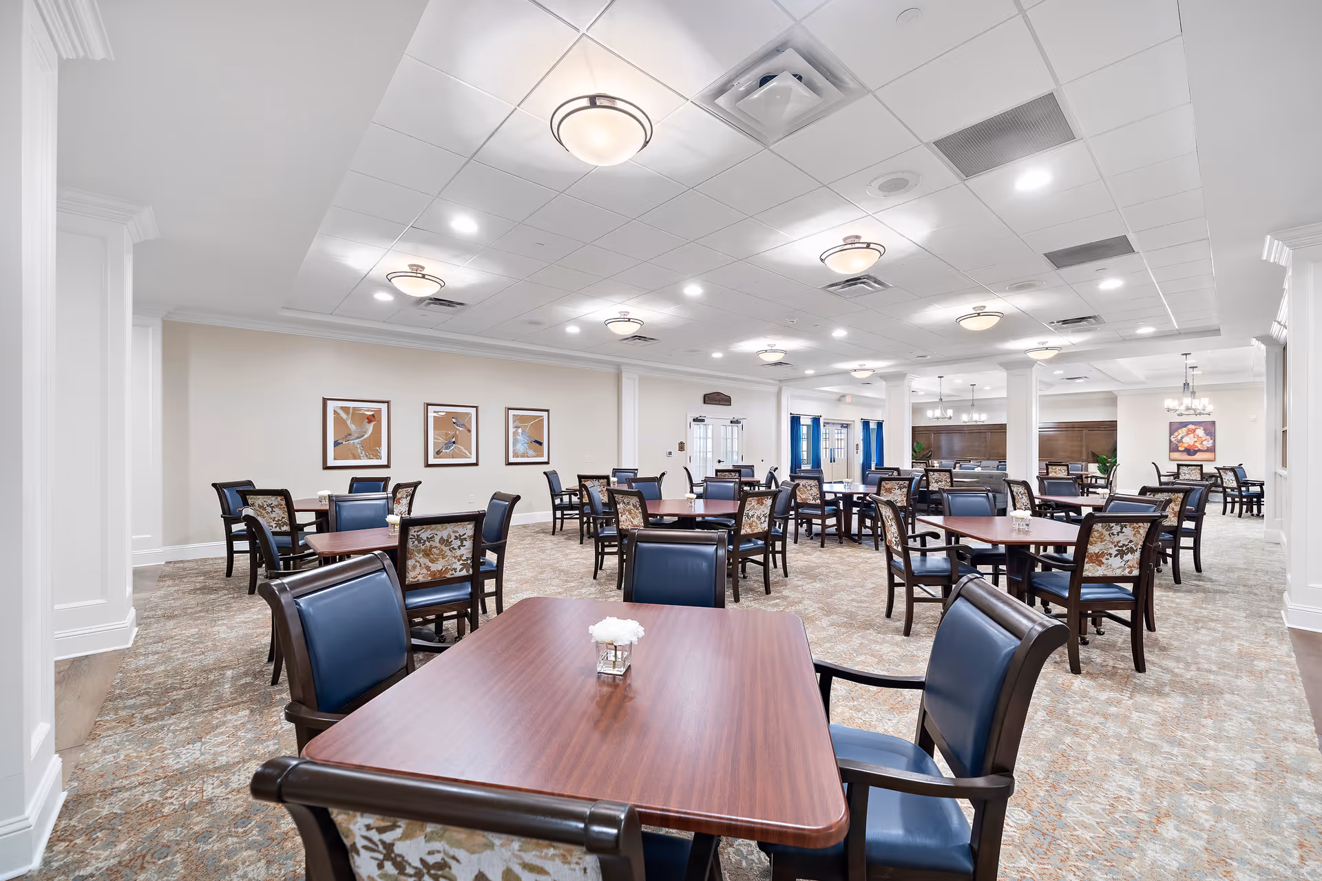 Spacious dining room with multiple wooden tables and upholstered chairs arranged under recessed ceiling lights.