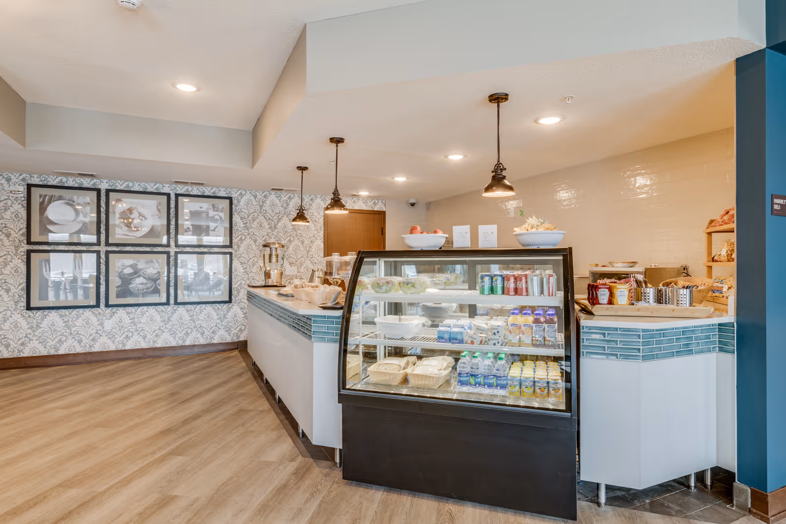 Interior view of a small deli or snack counter area with a glass display case filled with beverages and packaged food items. The counter has hanging pendant lights above it, and there are framed black and white photos on the patterned wallpapered wall to the left. The floor is wood-style, and the area appears clean and well-lit.