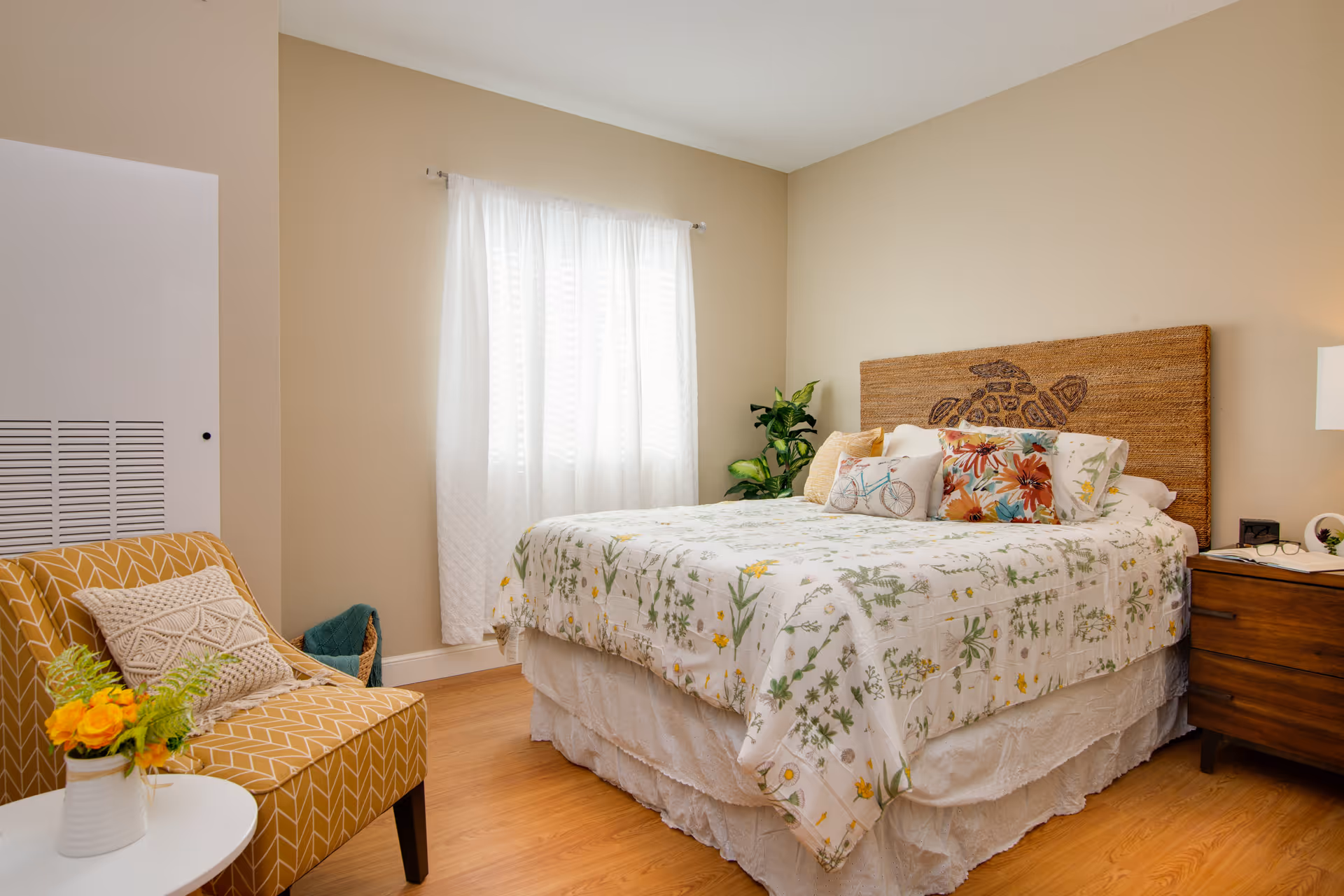 A cozy bedroom with a bed featuring a floral patterned bedspread and decorative pillows, including one with a bicycle design. The bed has a woven headboard with a turtle design. Next to the bed is a wooden nightstand with a lamp and some items on it. A yellow patterned armchair with a textured pillow is positioned near a small white round table holding a vase with yellow flowers. The room has light beige walls, a window with sheer white curtains, and wooden flooring.