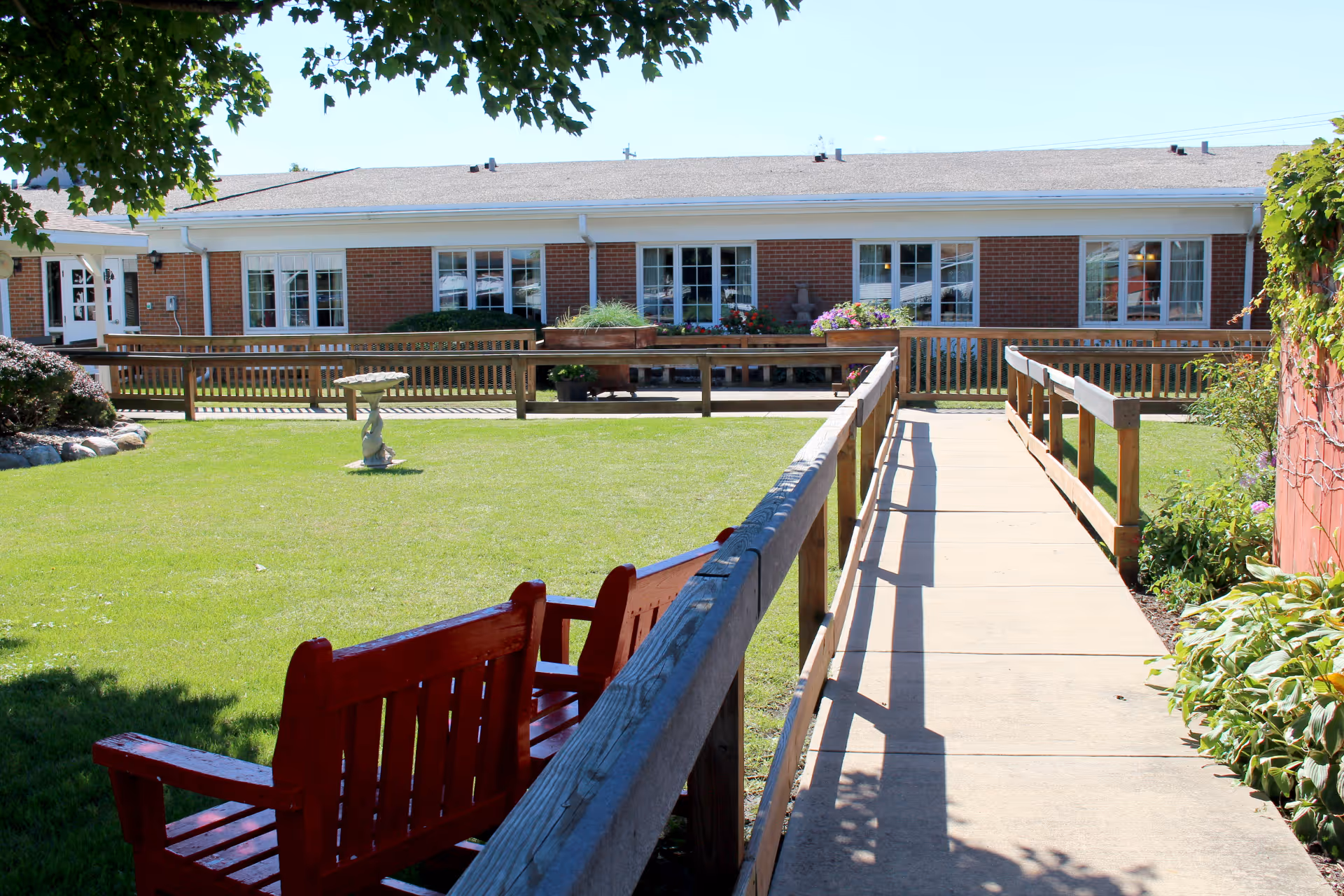 Outdoor view of a senior living facility with a concrete walkway bordered by wooden railings leading to a brick building with multiple windows. There is a well-maintained green lawn with a red wooden bench and some plants and flowers around the area.
