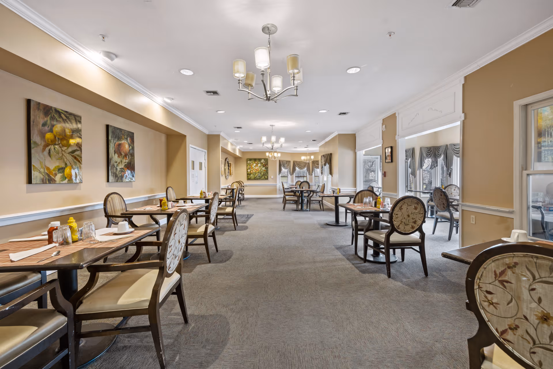 A spacious dining room with multiple tables and chairs arranged neatly. Each table is set with napkins, glasses, and condiments including mustard and ketchup. The room has beige walls adorned with framed paintings of fruit, and large windows with draped curtains allowing natural light to enter. Ceiling lights and chandeliers provide additional illumination.