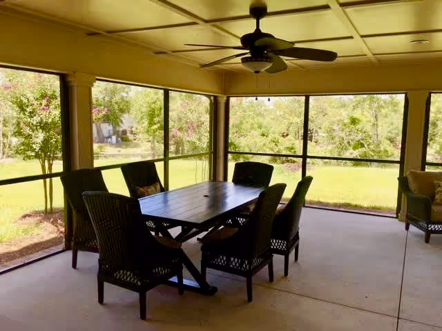 A screened-in patio area with a rectangular wooden table surrounded by six wicker chairs. The patio overlooks a green lawn with trees and bushes in the background.