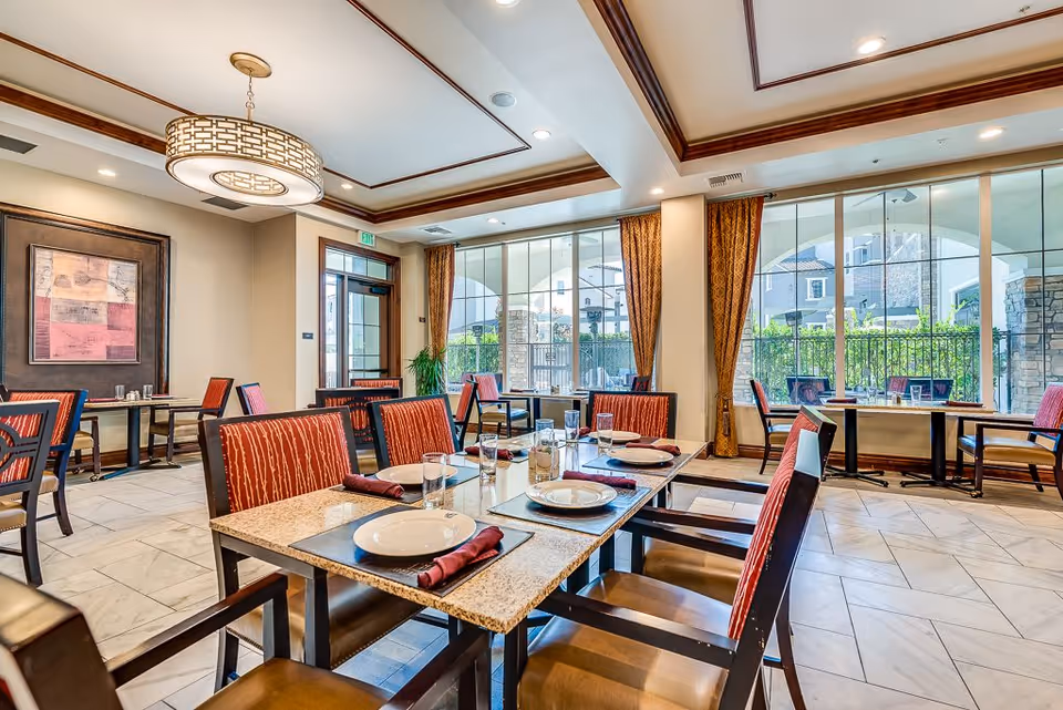 Bright and elegant dining room with multiple tables set with plates, glasses, and napkins. The room features large windows with brown curtains, allowing natural light to fill the space. There is a modern chandelier hanging from the ceiling and framed artwork on the wall.