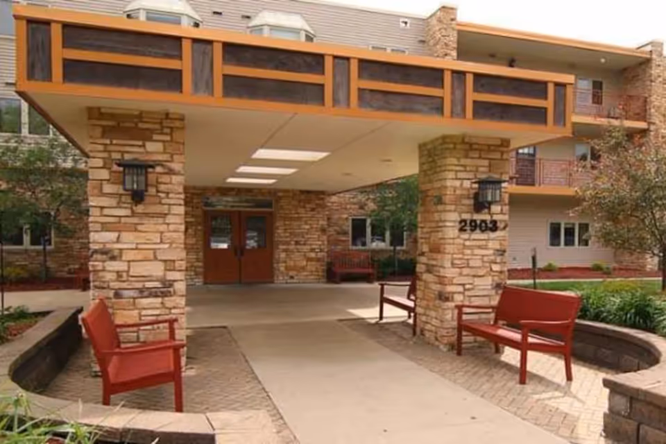 Entrance to a senior living facility with a covered drop-off area supported by stone pillars, red benches on either side, and a building with balconies and windows in the background.