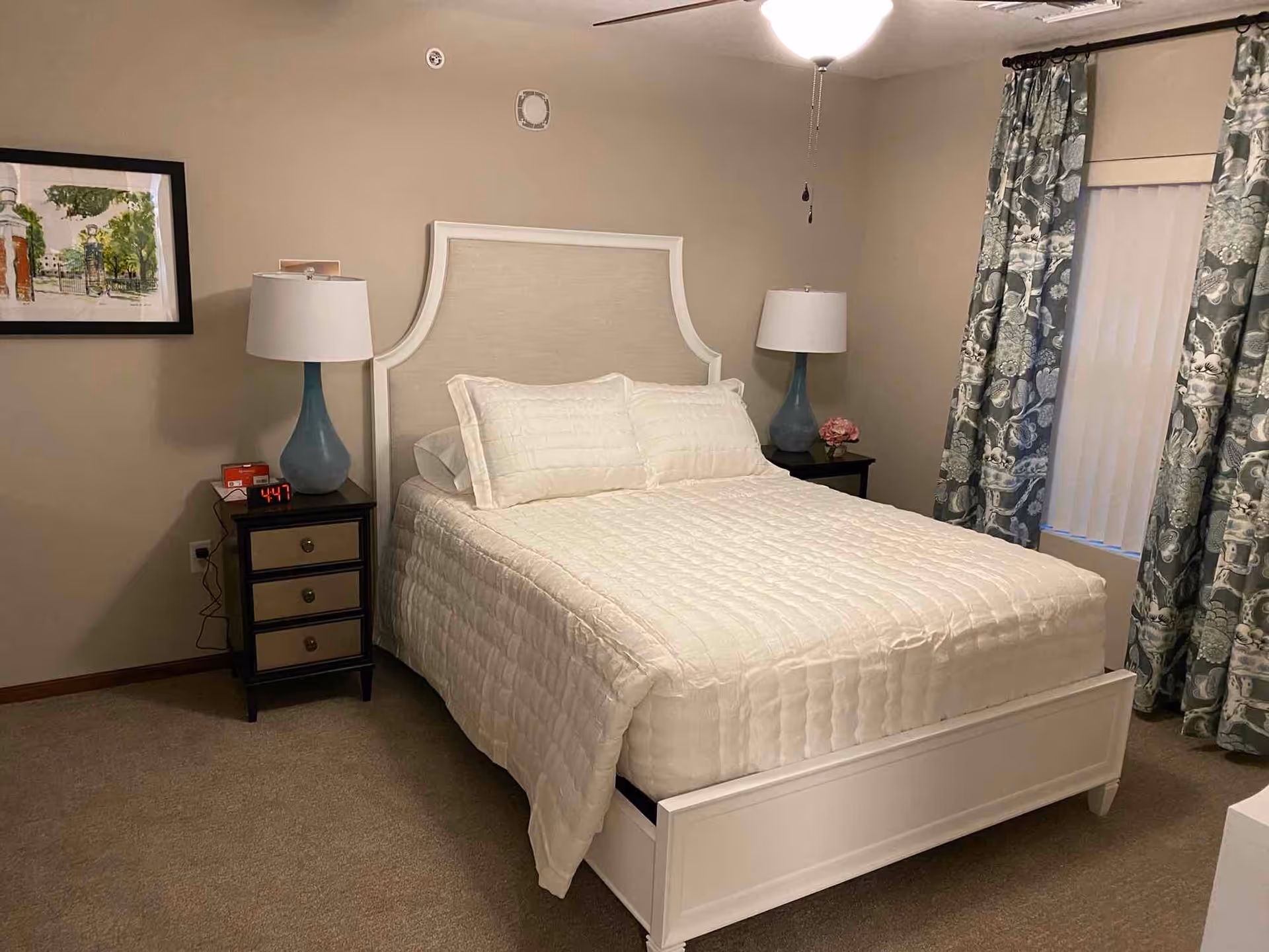 A neatly made bed with a white quilt and pillows in a bedroom. The bed has a white headboard and footboard. On either side of the bed are dark wooden nightstands with drawers, each holding a blue lamp with a white lampshade. One nightstand also has a digital alarm clock showing 4:47 and a small red box. There is a framed picture on the beige wall to the left and patterned blue and white curtains covering a window on the right side of the room. The floor is carpeted in a neutral color.