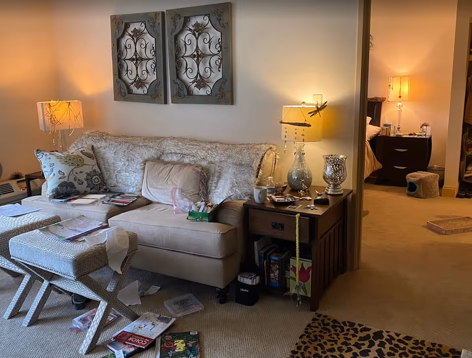 A cozy living room with a beige couch covered with a fluffy throw and decorative pillows. Two patterned ottomans are in front of the couch, with papers and books scattered on and around them. A wooden side table next to the couch holds a lamp, a cup, and various small items. The room is warmly lit by lamps, and a doorway leads to a bedroom with a nightstand, lamp, and pet furniture visible. The walls are decorated with two ornate metal wall hangings.