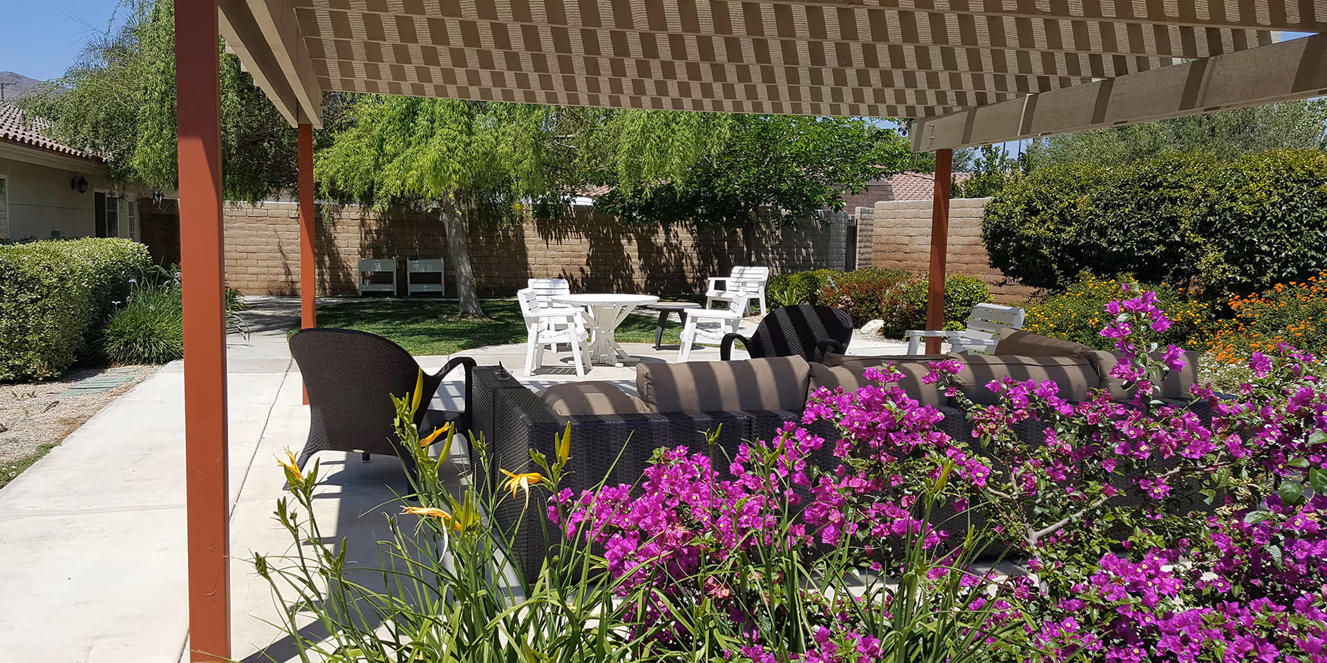 Outdoor seating area at The Gardens of Riverside featuring cushioned wicker chairs and a sofa under a shaded pergola, surrounded by vibrant purple flowers and greenery with a concrete walkway and additional white chairs and tables in the background.