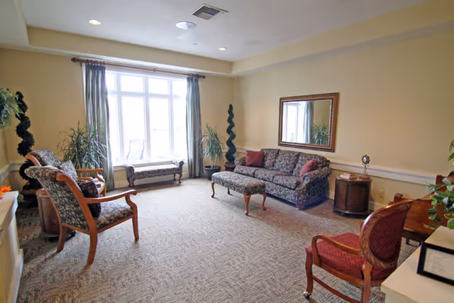 A well-lit living room in Cedar Ridge Senior Living featuring a large window with curtains, a patterned sofa with matching ottoman, two armchairs, a bench by the window, a side table with a clock, a large mirror on the wall, and several potted plants.