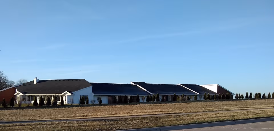 Single-story long white building with a dark roof set behind a grassy field under a clear blue sky.
