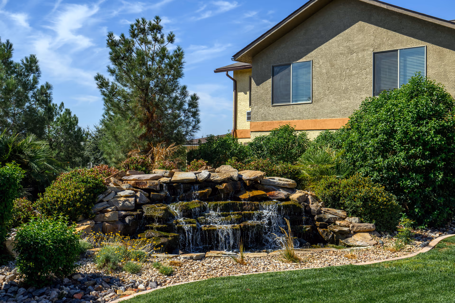 Outdoor garden area at The Meadows at Escalante featuring a small cascading rock waterfall surrounded by green bushes, trees, and a well-maintained lawn, with part of a beige building with two windows in the background under a blue sky with wispy clouds.