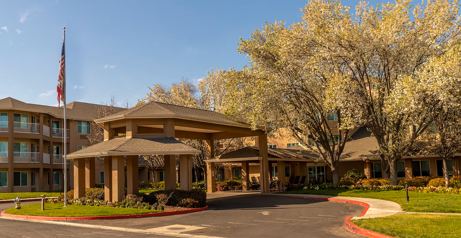 Exterior view of Sierra Hills - A Provincial Senior Living Community, showing a multi-story building with balconies, a covered entrance driveway, blooming trees, and a flagpole with the American and California flags.