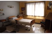 A nursing home bedroom with a hospital bed positioned near a large window with sheer curtains. The room includes a bedside table, a chair, a lamp, and a small table with a television on it. The walls are painted in light and warm tones, and the floor has a patterned rug.