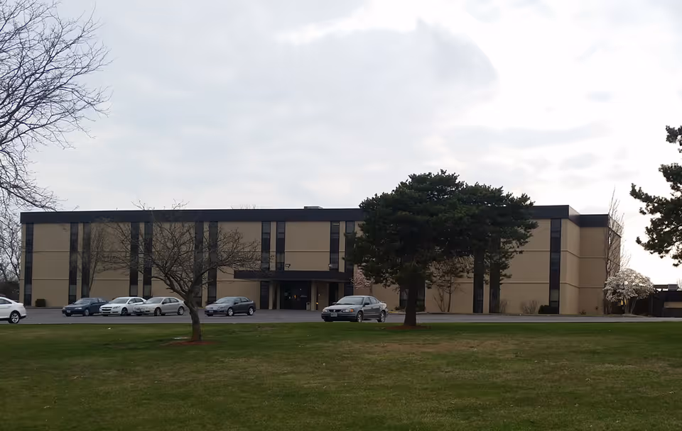 Exterior view of a three-story beige building with vertical dark window panels, a parking lot with several cars in front, and a grassy area with a few trees in the foreground under a cloudy sky.
