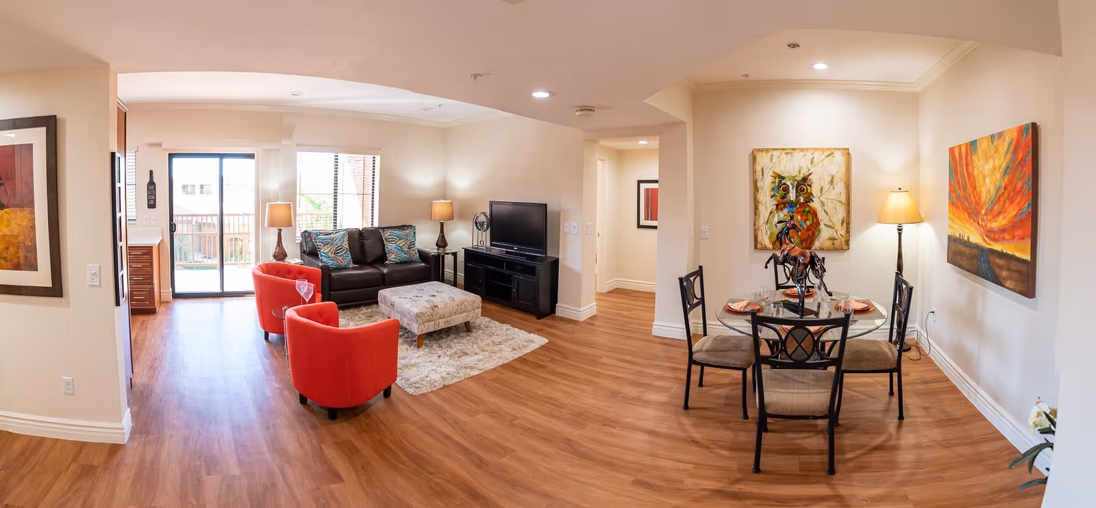 A bright and spacious living and dining area with wooden flooring. The living area features a black leather sofa with patterned cushions, two red armchairs, a patterned ottoman on a fluffy rug, and a TV on a black stand. The dining area has a round glass table with four chairs, set with plates and glasses, and decorated with a horse sculpture. The walls are adorned with colorful paintings, and there are several lamps providing warm lighting. A sliding glass door leads to an outdoor balcony.