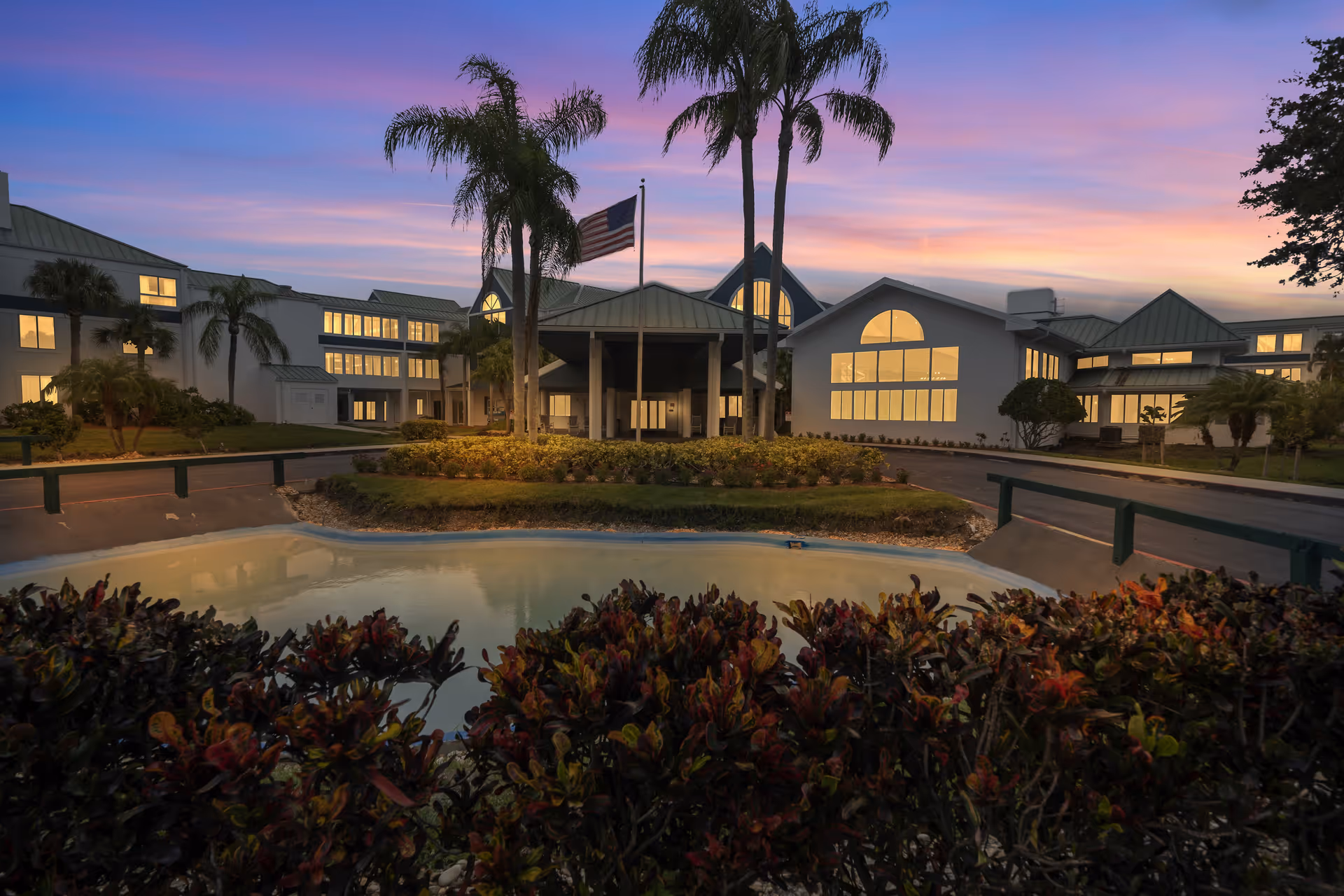 Twilight view of the front entrance of a senior living facility with palm trees, an American flag, a landscaped pond, and illuminated windows.