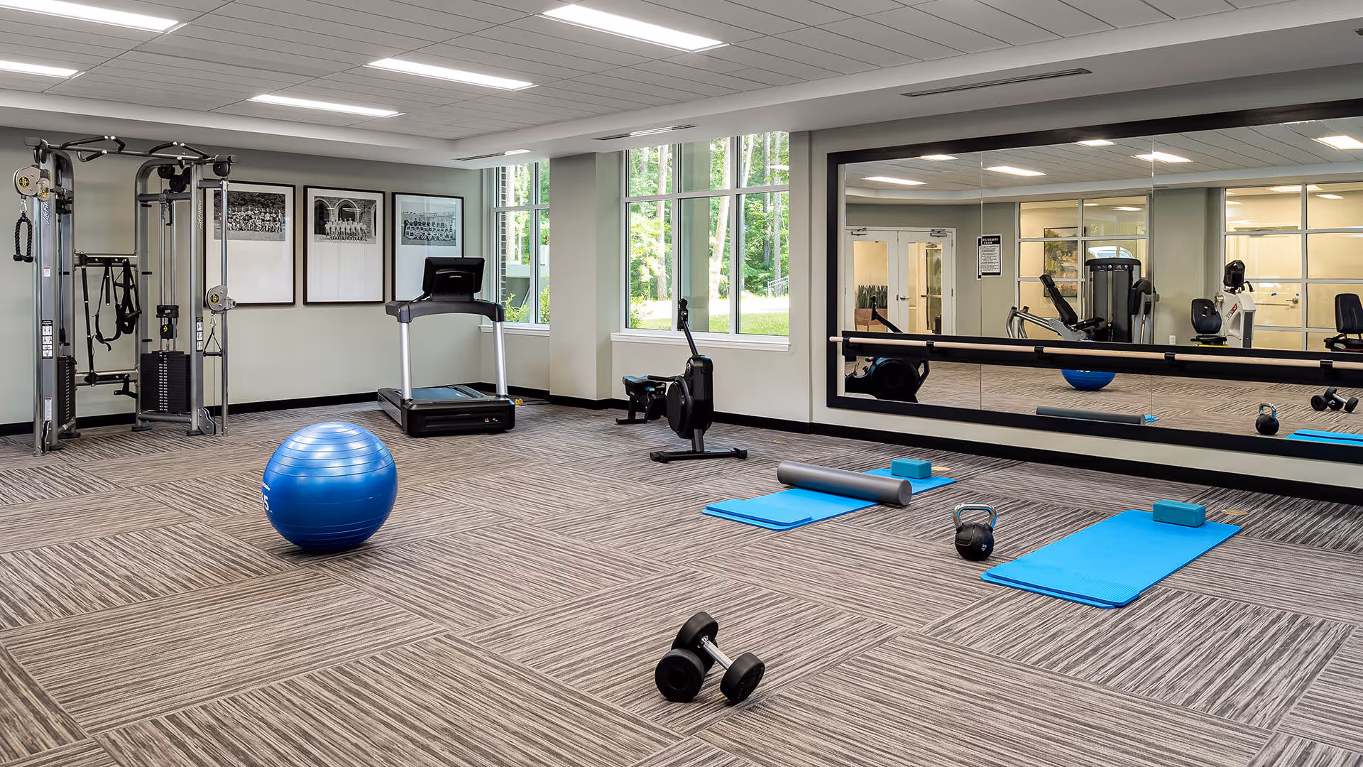A fitness room with exercise equipment including a treadmill, rowing machine, weight machine, dumbbells, kettlebell, yoga mats, foam roller, yoga blocks, and a large blue exercise ball. The room has large windows letting in natural light and a wall-sized mirror reflecting the equipment.