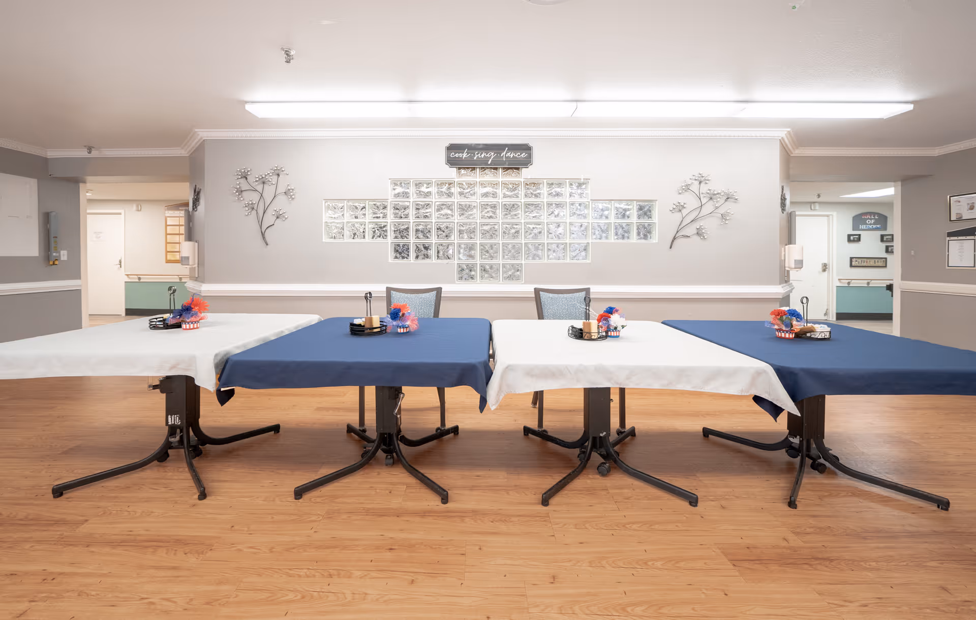 A dining area with four tables covered in alternating blue and white tablecloths, each decorated with small patriotic centerpieces. The room has light-colored walls with decorative metal wall art and a sign that reads 'cook. serve. dance.' The floor is wood, and there are chairs placed behind the tables. The space appears clean and well-lit.