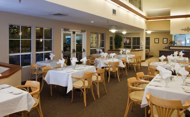 A dining room in a senior living facility with multiple tables covered in white tablecloths, each set with plates, glasses, and folded napkins. Wooden chairs surround the tables, and large windows and double doors are visible along one wall. The room is well-lit with ceiling lights and has a warm, inviting atmosphere.