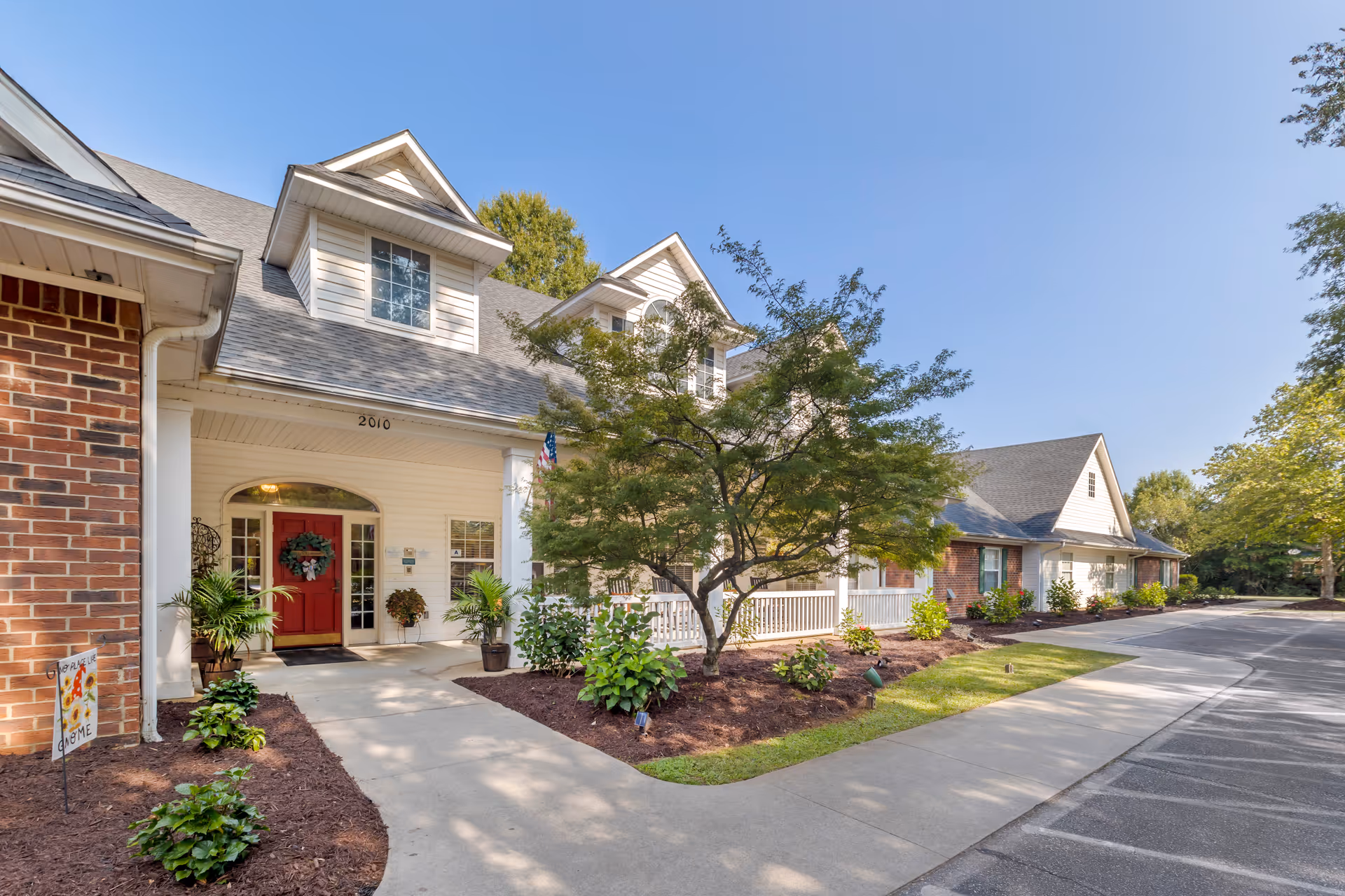 Exterior front entrance of a senior living building with a red door, porch, landscaping and a driveway under a clear sky.