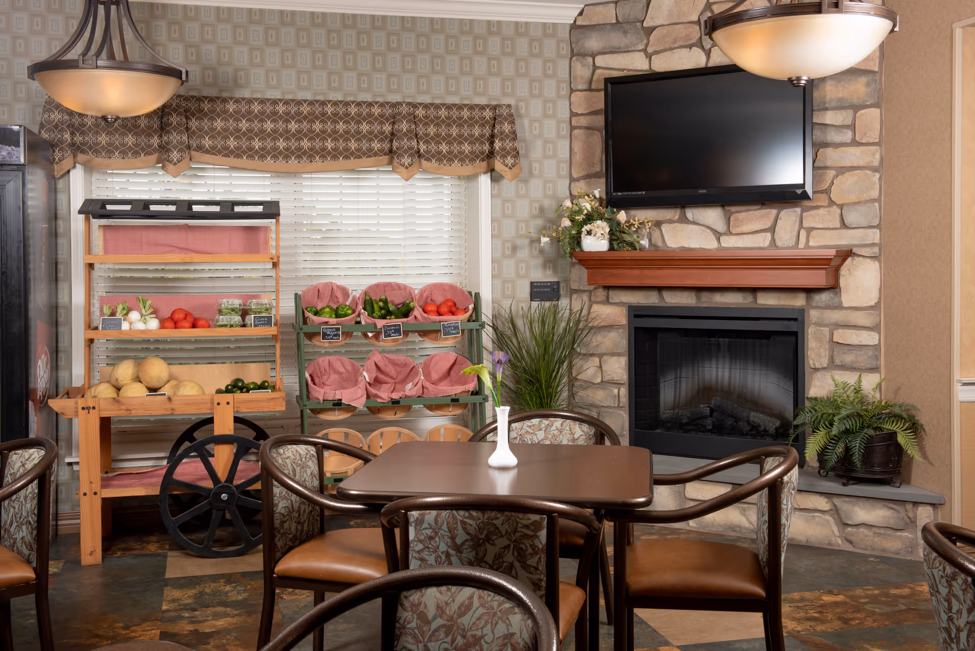 A cozy dining area with a square table and four chairs featuring floral upholstery. Behind the table, there is a stone fireplace with a wooden mantel and a flat-screen TV mounted above it. To the left, there are wooden shelves and baskets holding fresh vegetables like tomatoes, cucumbers, and melons. The window behind the shelves has white blinds and a patterned valance. The room is warmly lit by hanging light fixtures and decorated with potted plants.