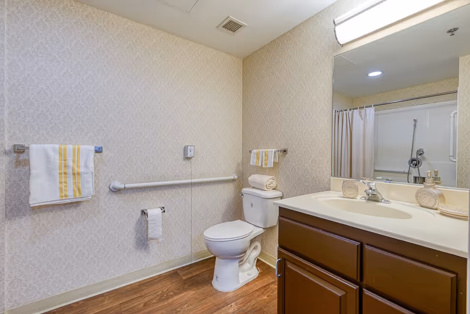 A clean bathroom with a toilet, a vanity with a sink and mirror, a towel rack with a white towel with yellow stripes, a toilet paper holder, and a shower with a curtain. The walls have a subtle patterned wallpaper and the floor is wood.