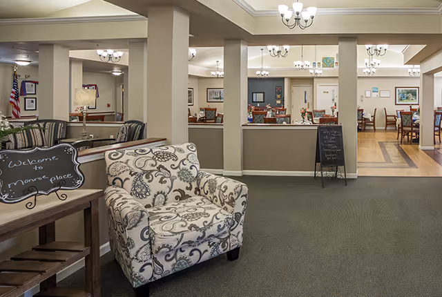Interior view of a senior living facility common area with a patterned armchair in the foreground, a small chalkboard sign on a wooden table, and multiple dining tables and chairs in the background under chandeliers.