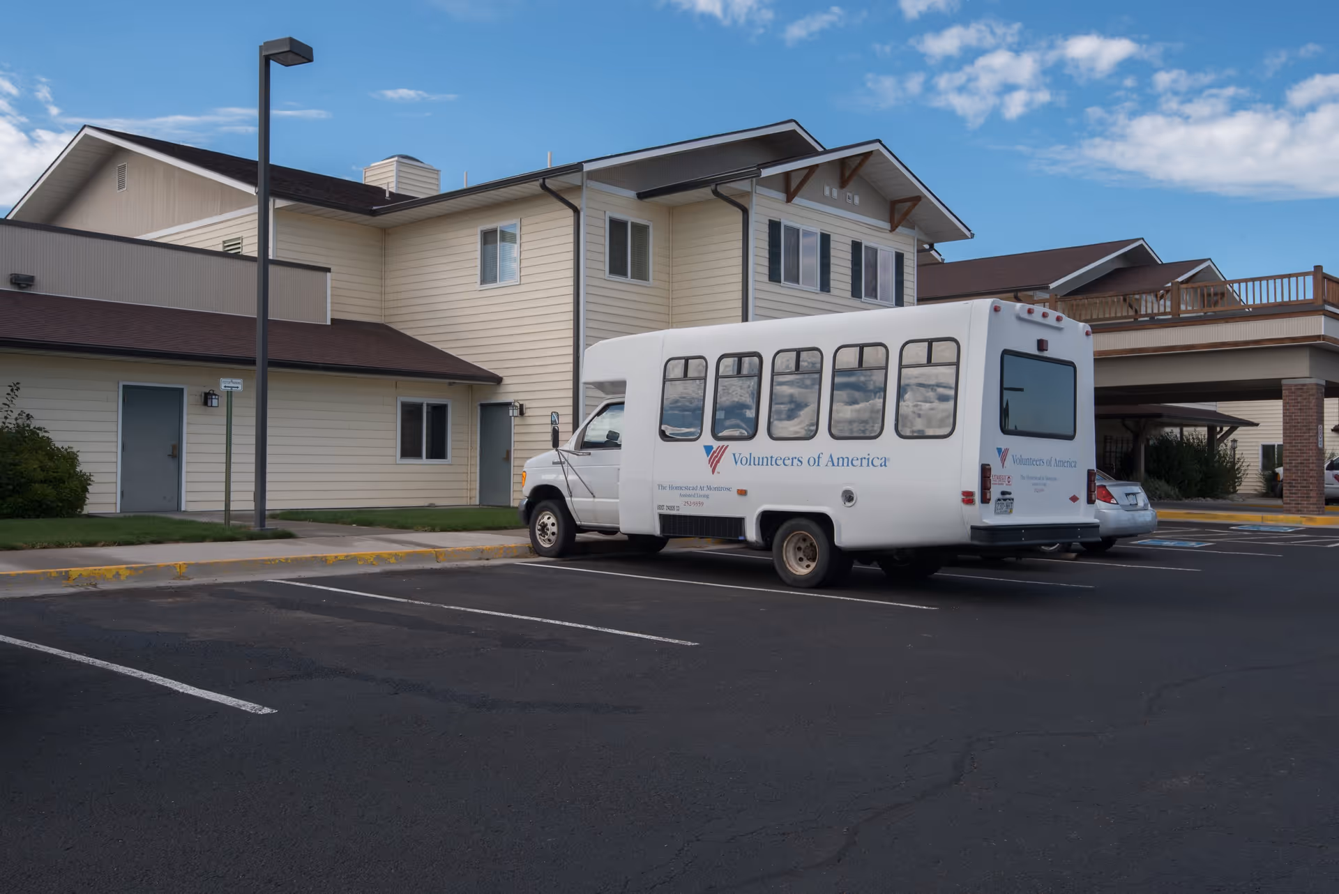 Exterior view of The Homestead At Montrose facility with a white Volunteers of America shuttle bus parked in the parking lot under a clear blue sky.