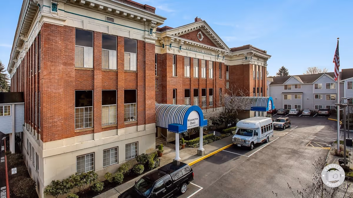 Exterior view of a large brick building with white trim labeled Washington School, featuring blue and white striped awnings over the entrances marked Washington Oakes. Several vehicles, including a white van and a black truck, are parked in the adjacent parking lot. An American flag is visible on a flagpole to the right.