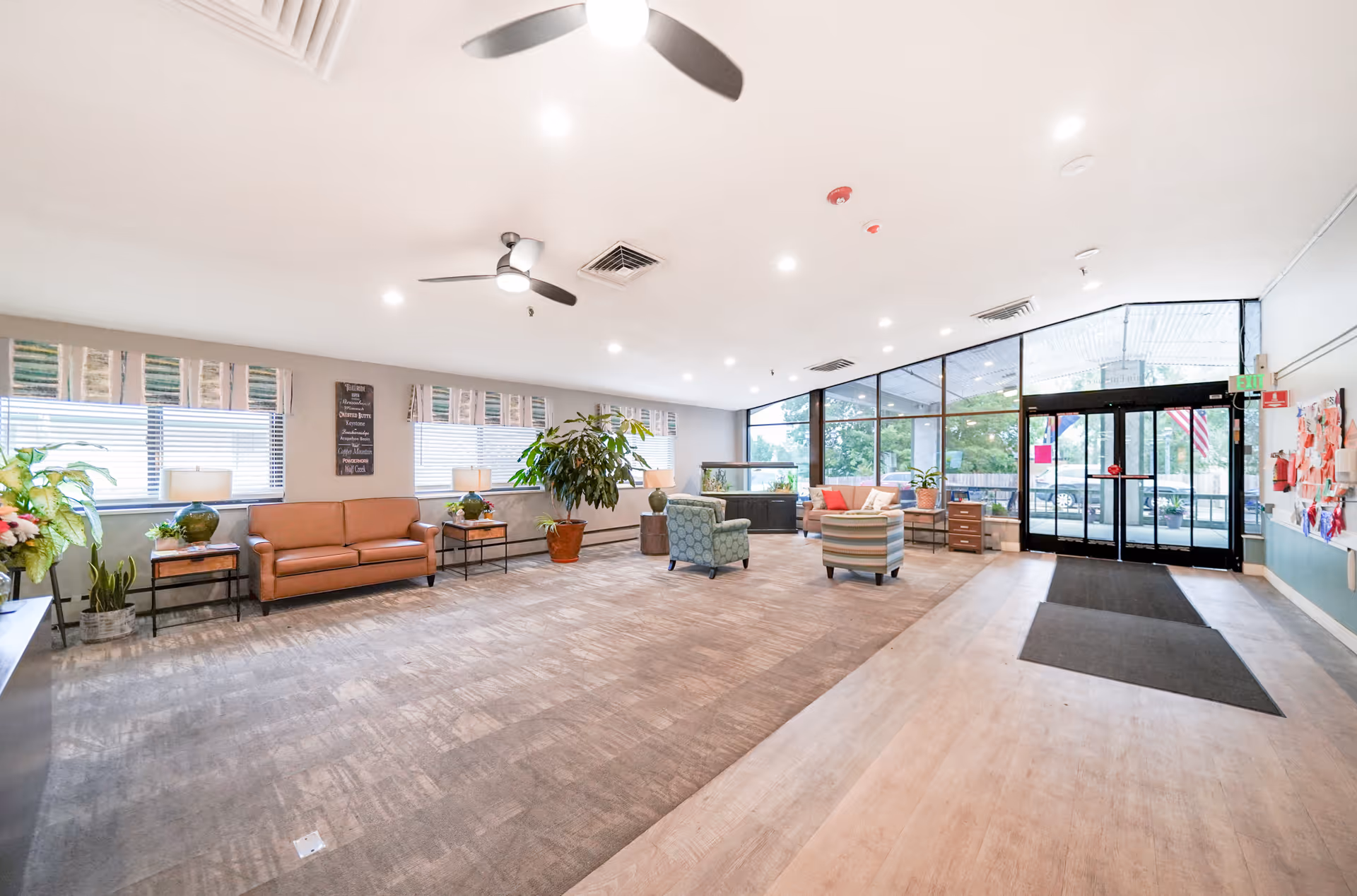 A spacious and well-lit common area with large windows and glass doors letting in natural light. The room features a mix of seating including a brown leather sofa, patterned armchairs, side tables with lamps, and several potted plants. Ceiling fans and recessed lighting are visible on the white ceiling. The entrance door has an exit sign above it and there is a bulletin board on the right wall.